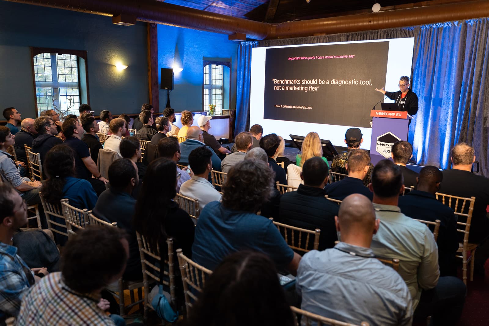 Isaac standing at a lectern in front of a room of seated people gesturing to a slide that reads '"Benchmarks should be a diagnostic tool, not a marketing flex" - Isaac Z. Schlueter, NodeConf EU, 2024'