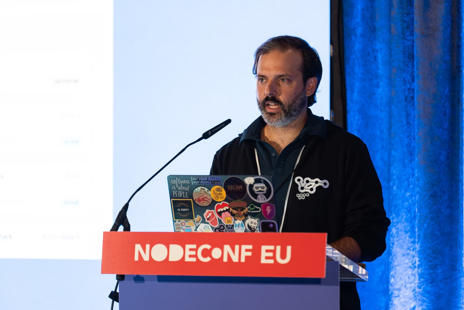 Luke standing at a lectern with the words "NodeConf EU" on the front with a sticker covered laptop in front of him.