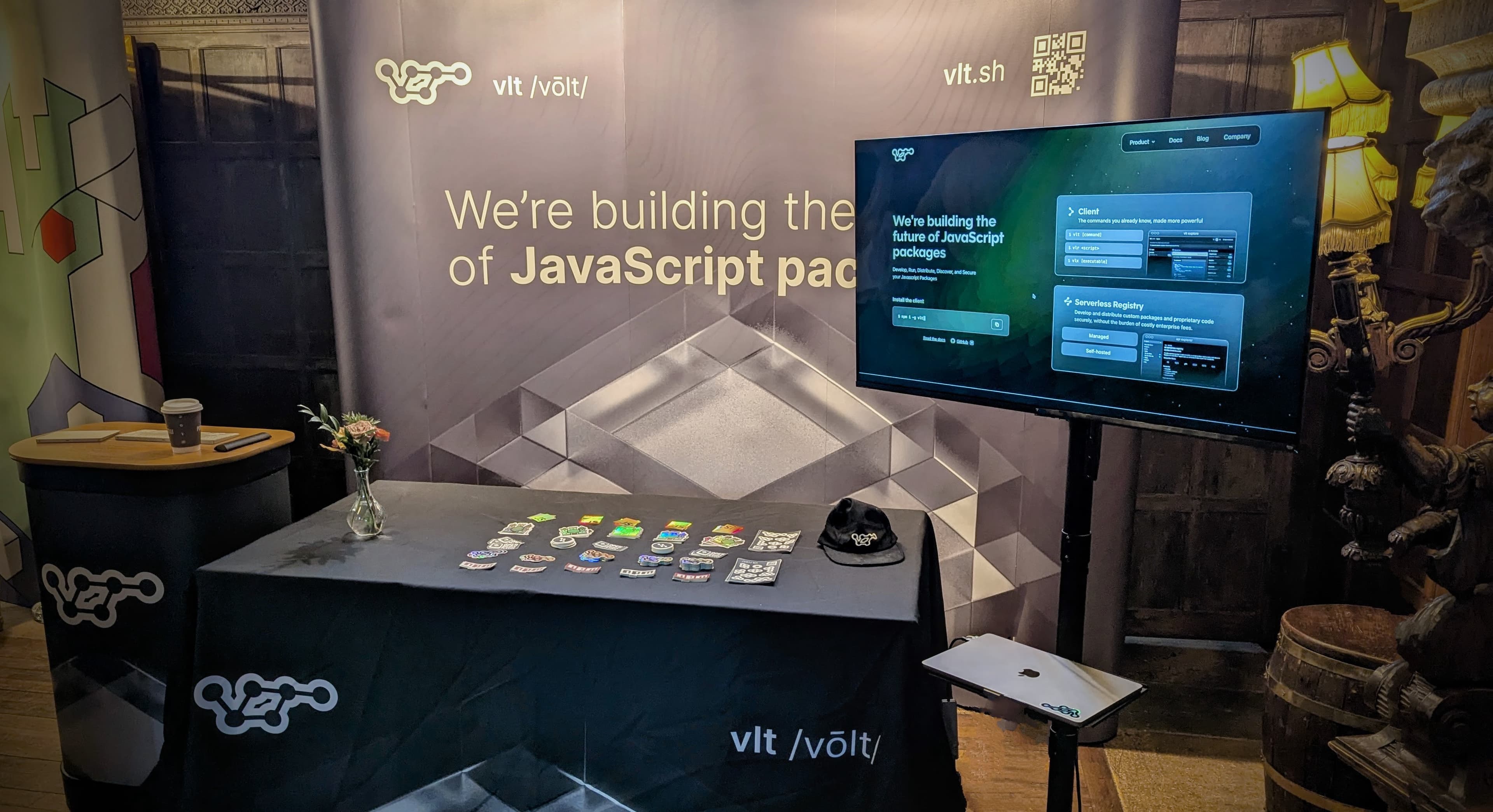 A table with rows of neatly organized stickers. Behind the table is a booth that reads "Builing the future of JavaScript packages". There is a TV to right right showing the vlt.sh homepage.