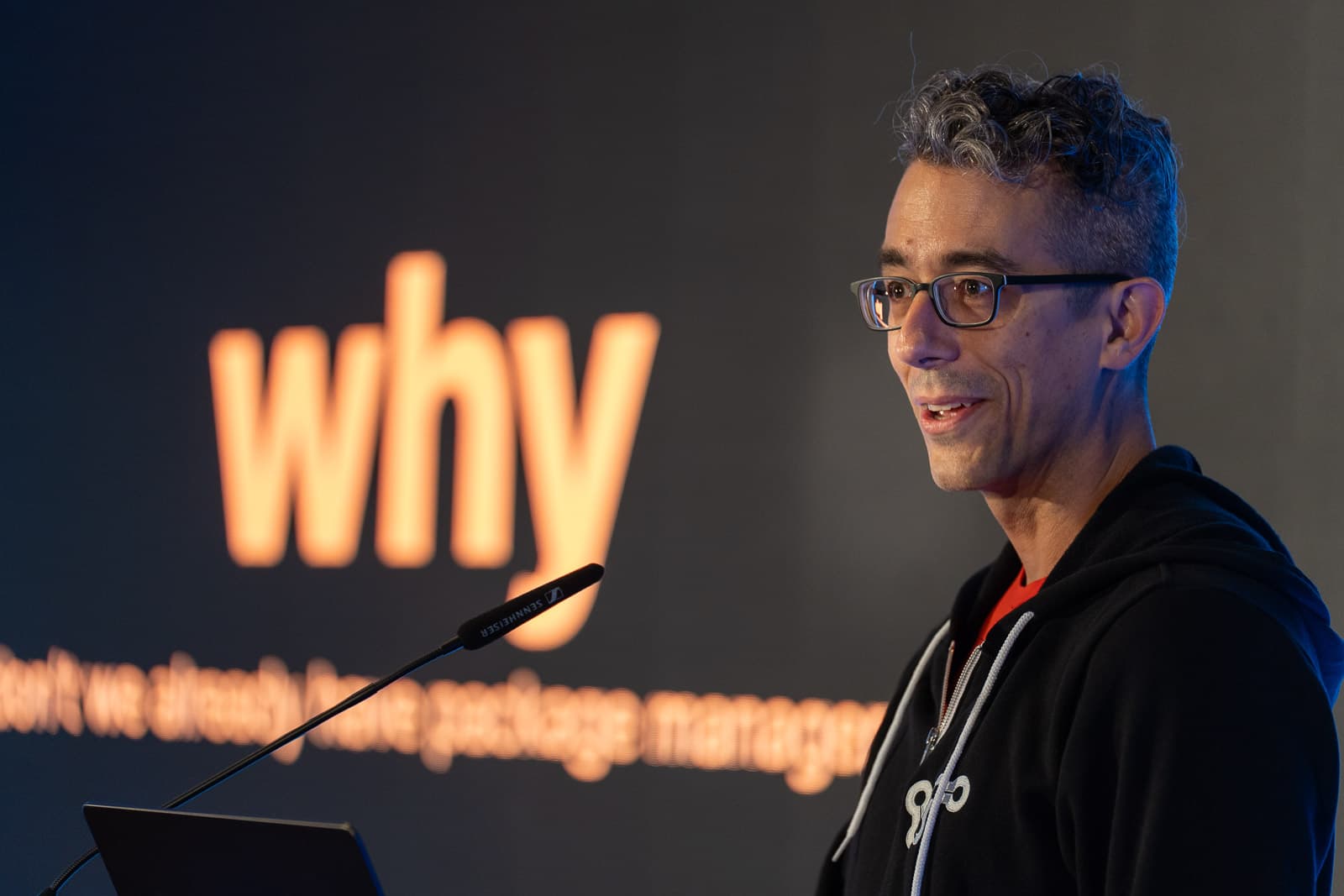 Isaac wearing a black hoodie and smiling in front of a slightly out-of-focus slide with the word "why" on it.